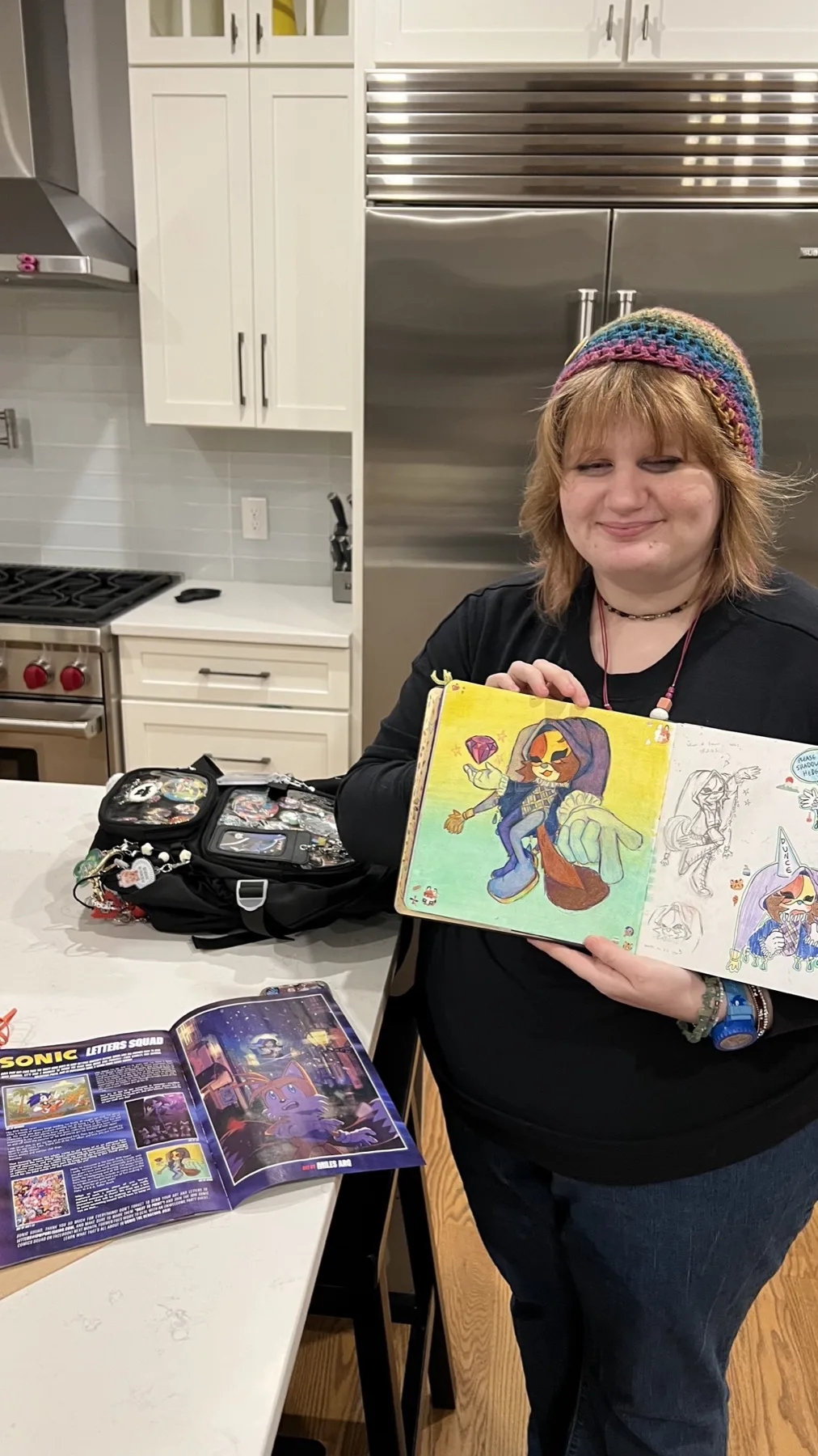 Ophir smiling in a kitchen, holding her sketchbook open beside the comic where her artwork was published